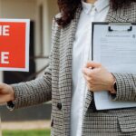 a-real-estate-agent-holding-a-home-for-sale-sign-and-clipboard-outside-a-property