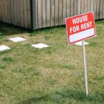 a-red-house-for-rent-sign-stands-on-a-grassy-lawn-beside-a-wooden-house-exterior