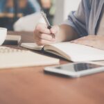 a-woman-writes-in-a-notebook-at-a-cafe-table-with-a-coffee-and-smartphone-nearby