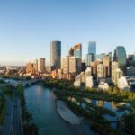 A wide-angle view of a city's skyline with tall buildings, a river, and green spaces on a clear day. A bridge crosses the river on the left side of the image.