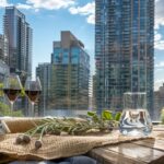 A cityscape view from a rooftop garden at 9Hundred Signature Residences features a wooden tray with two glasses of red wine, a water glass, a bottle, and greenery, set against tall modern buildings.