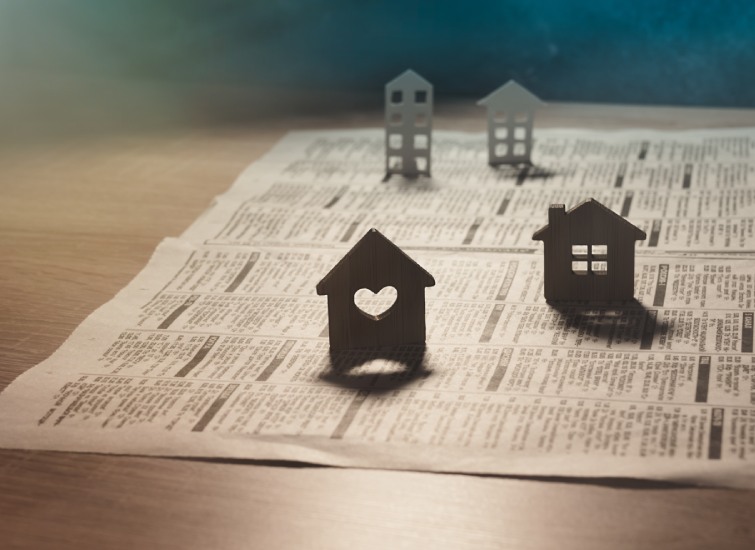 Three wooden house-shaped cutouts on top of a newspaper, casting shadows on the surface, with a warm light source on one side.