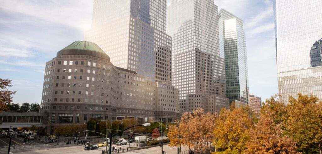 Cityscape with tall skyscrapers, a building featuring a green dome, and autumn trees in the foreground. A street with traffic and pedestrians is visible at the bottom.