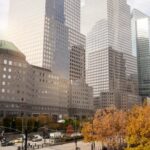 Cityscape with tall skyscrapers, a building featuring a green dome, and autumn trees in the foreground. A street with traffic and pedestrians is visible at the bottom.