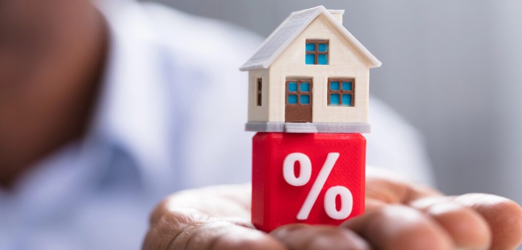 A person holds a small model house that is placed on top of a red cube with a white percentage symbol.