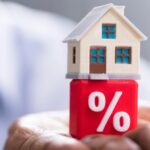 A person holds a small model house that is placed on top of a red cube with a white percentage symbol.