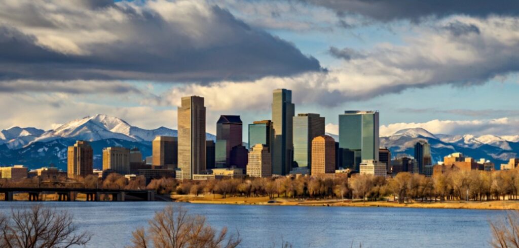 A city skyline with tall buildings in the foreground, snowy mountains in the background, and a lake with a bridge in the foreground.