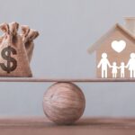 A bag with a dollar sign and a wooden house with a family icon are balanced on a wooden seesaw, positioned atop a wooden sphere.