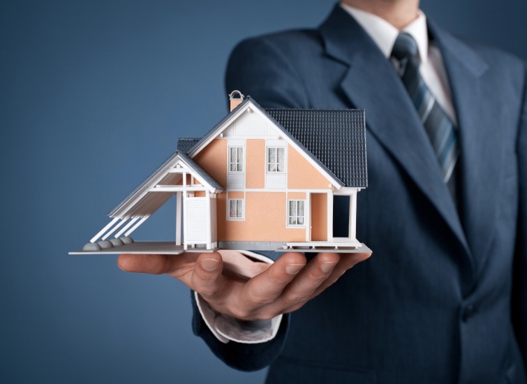 A person in a suit holding a small model of a house with a blue roof, white exterior, and attached carport.