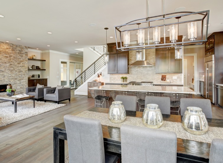 Modern open-concept kitchen and living area with a dining table in the foreground, sleek lighting fixtures, stainless steel appliances, dark wood cabinetry, and contemporary stone accents on the far wall.