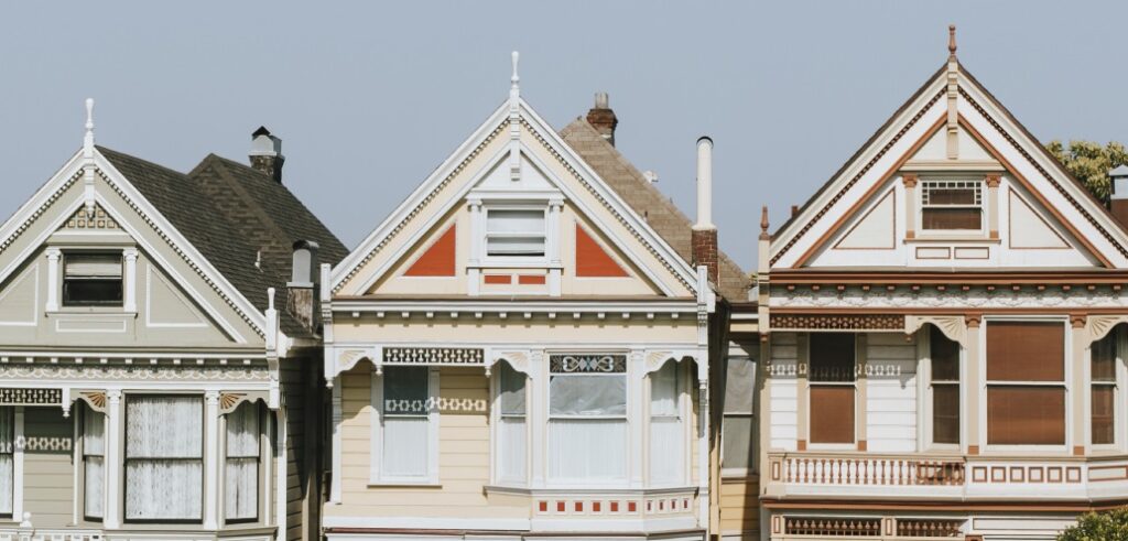 Three adjoining Victorian-style houses with ornate trim, each painted in different pastel colors. The houses have pointed roofs and large front windows.