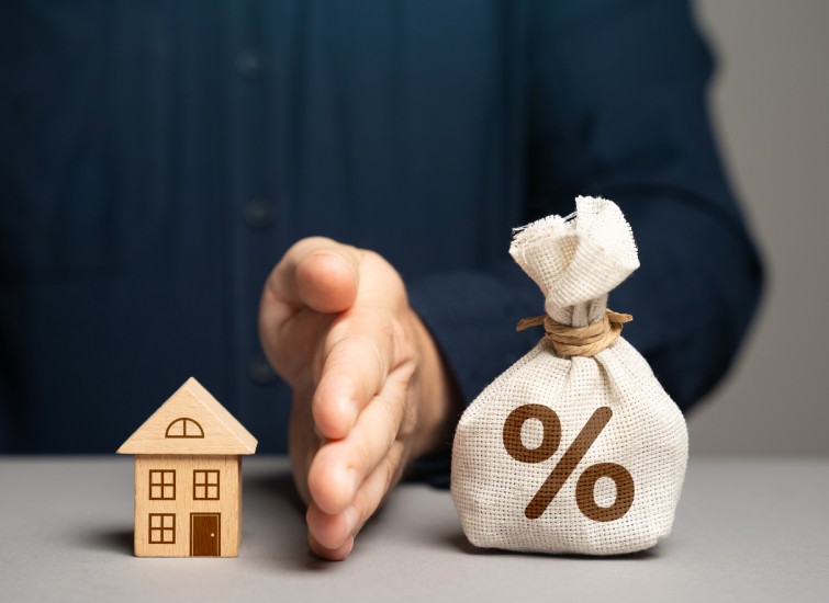 A man's hand holding a bag of money and a model of a house, symbolizing the real estate market.