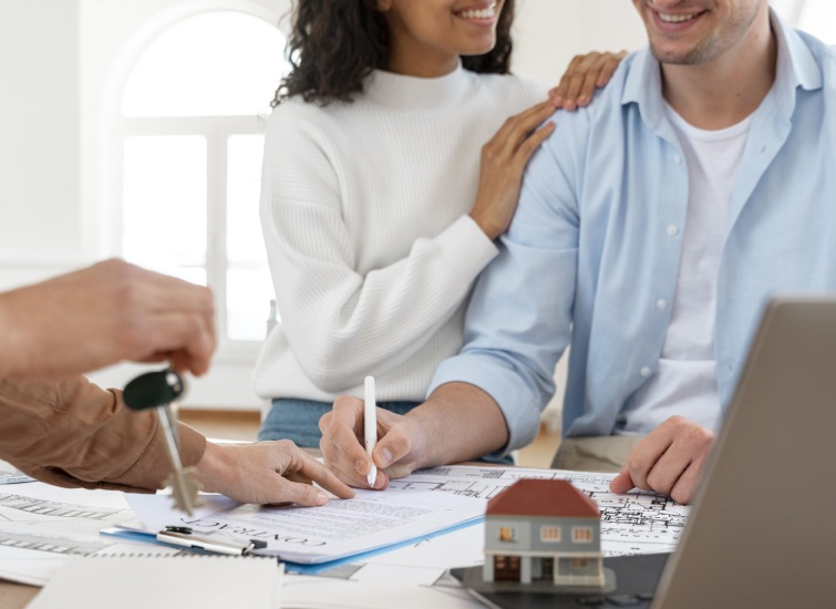 A couple signs documents while another person hands over a set of keys. A small model of a house is visible on the table beside them.