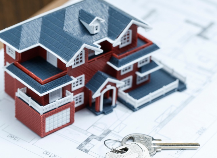 A miniature model house with blue and red colors is placed on architectural blueprints, with a set of silver keys in the foreground.