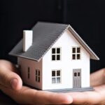 Person holding a small model of a white house with both hands.